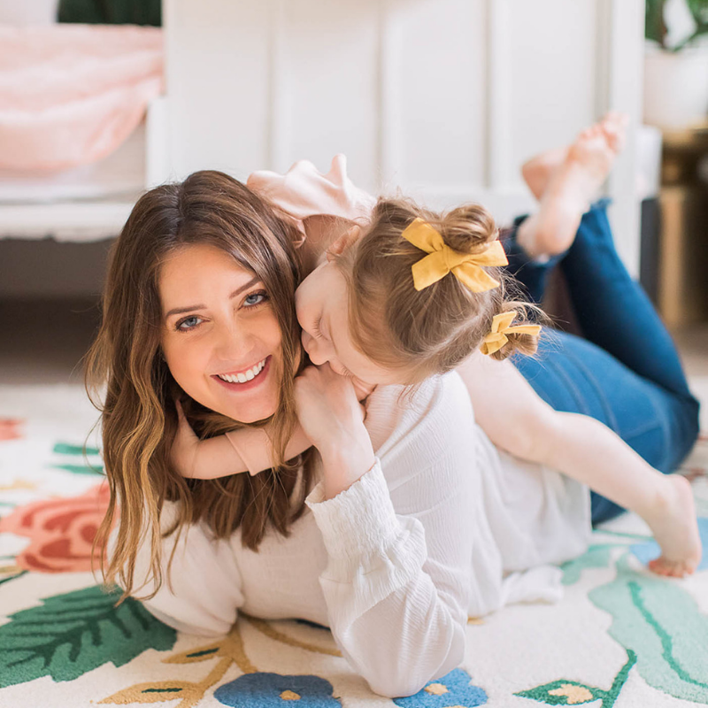 Woman lying on a colorful floral rug smiling while a young child with yellow hair bows cuddles on her back
