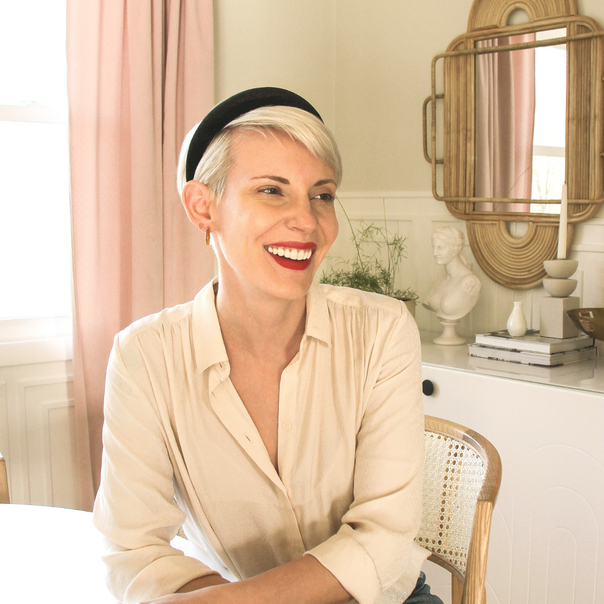 Smiling woman seated at a table in a light-filled room with pink curtains, rattan mirror, and decorative bust