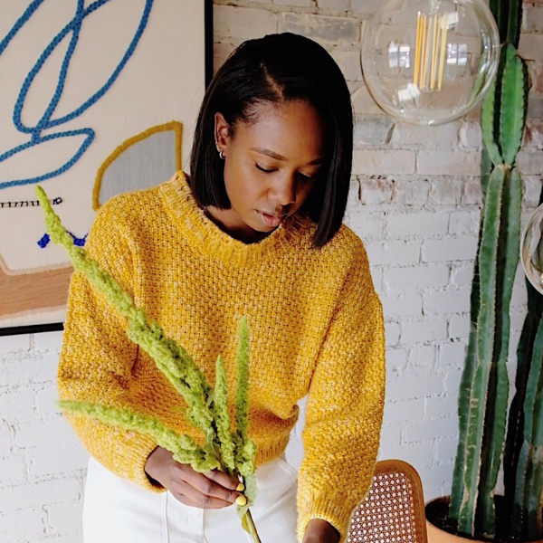 Elena Lohse arranging green stems in a sunlit living room with white brick wall, pendant glass lights, and a tall cactus