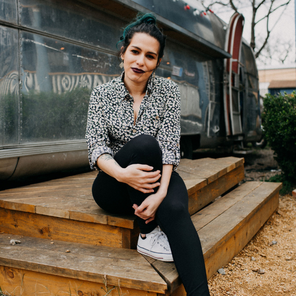 Woman with teal hair sitting on wooden steps in front of a silver trailer, wearing a floral blouse, black leggings, and white sneakers