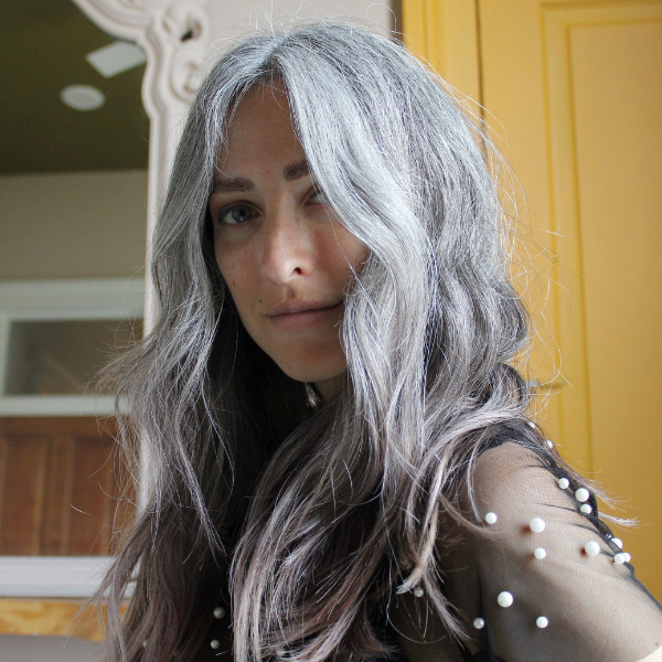 Woman with long silver hair looking at camera, wearing sheer black top with pearls in front of a yellow door