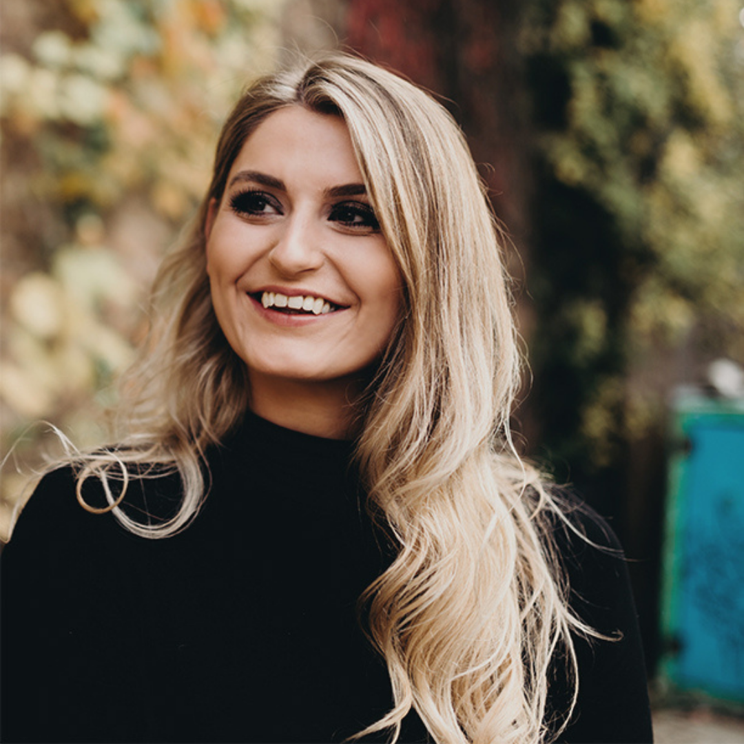 Smiling woman outdoors with long blonde hair wearing a black turtleneck, blurred garden background
