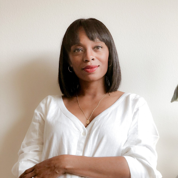 Portrait of a woman standing with arms crossed in a white blouse against a plain light wall, soft natural light