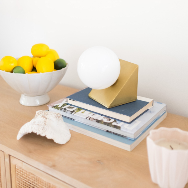 Brass geometric table lamp with white globe on stacked books beside a bowl of lemons on a light wood console