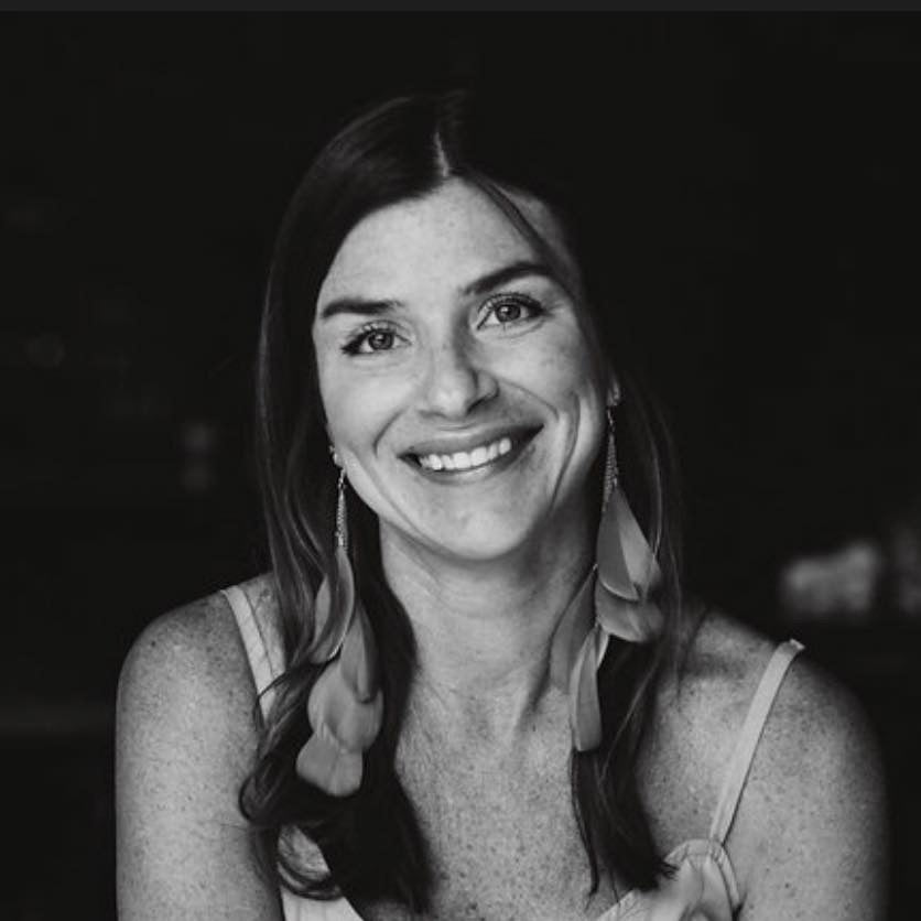 Head-and-shoulders portrait of a smiling woman wearing long feather earrings against a dark background