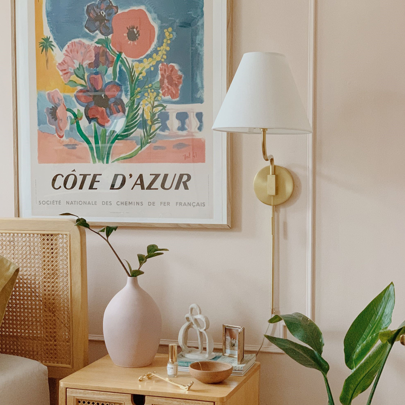 Bedside table styled with pink vase, wooden bowl and sculptural decor under a framed CÔTE D’AZUR poster with a brass wall sconce and houseplants