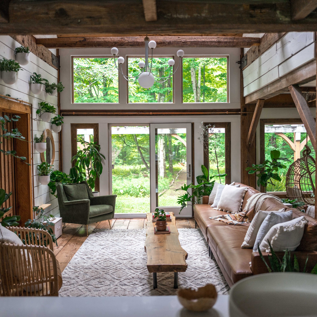 Cozy rustic living room with leather sofa, green armchair, live-edge wood coffee table, chandelier, and large windows overlooking a green forest