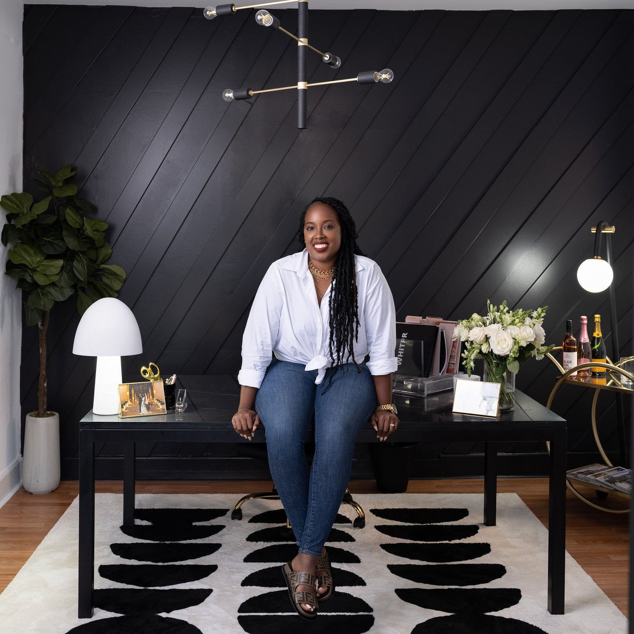 Dreena Whitfield sitting on a desk in her new office, smiling in front of a black diagonal paneled wall with lamp, flowers, and bottles