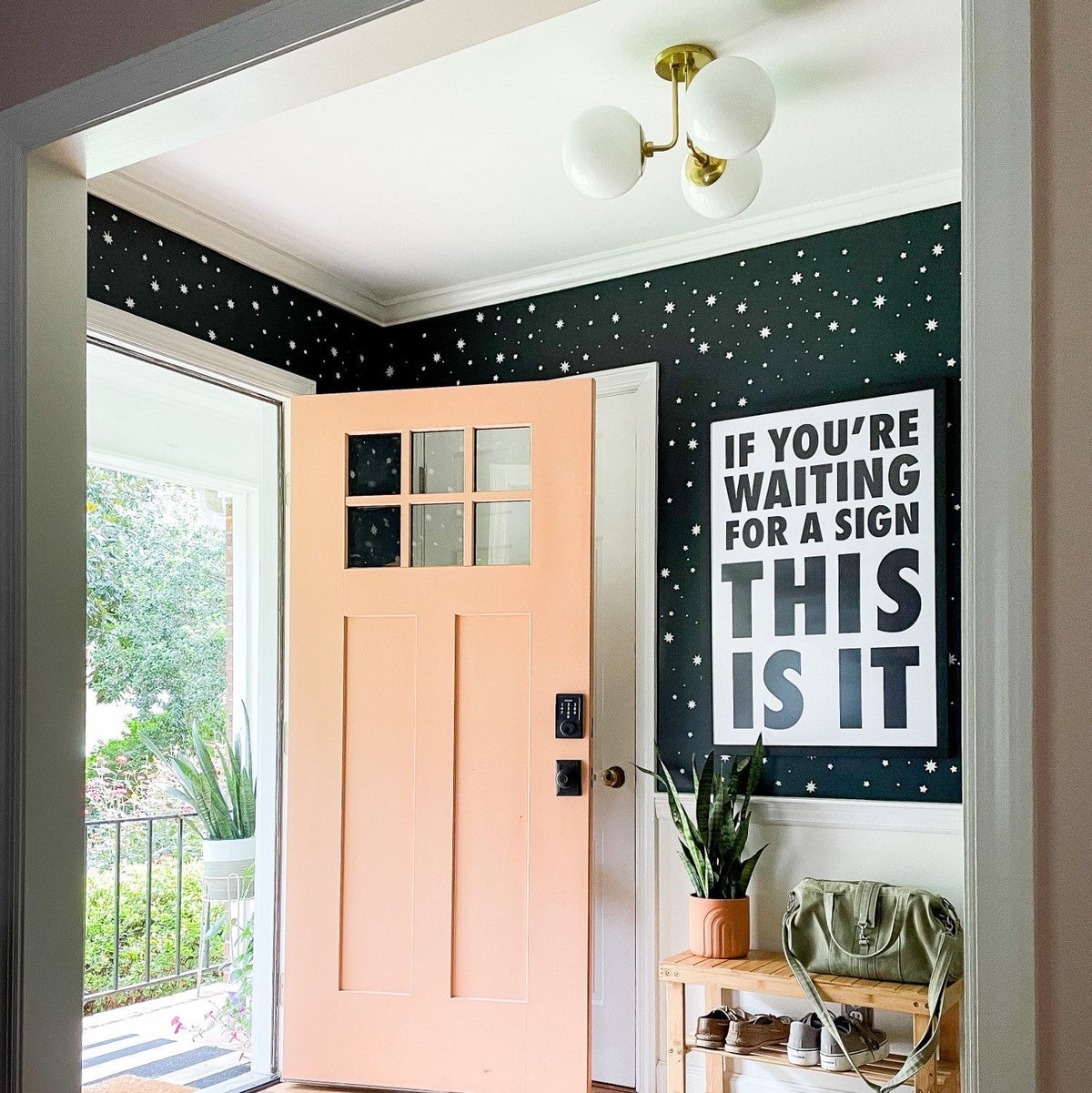Open peach front door revealing entryway with dark star-patterned wallpaper, globe ceiling light, potted plant, bench, and poster reading “IF YOU’RE WAITING FOR A SIGN THIS IS IT”