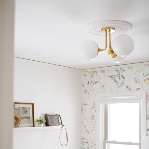 Brass three-globe flush-mount ceiling light over a bright bedroom corner with floral wallpaper, window, and a shelf with framed art and plant