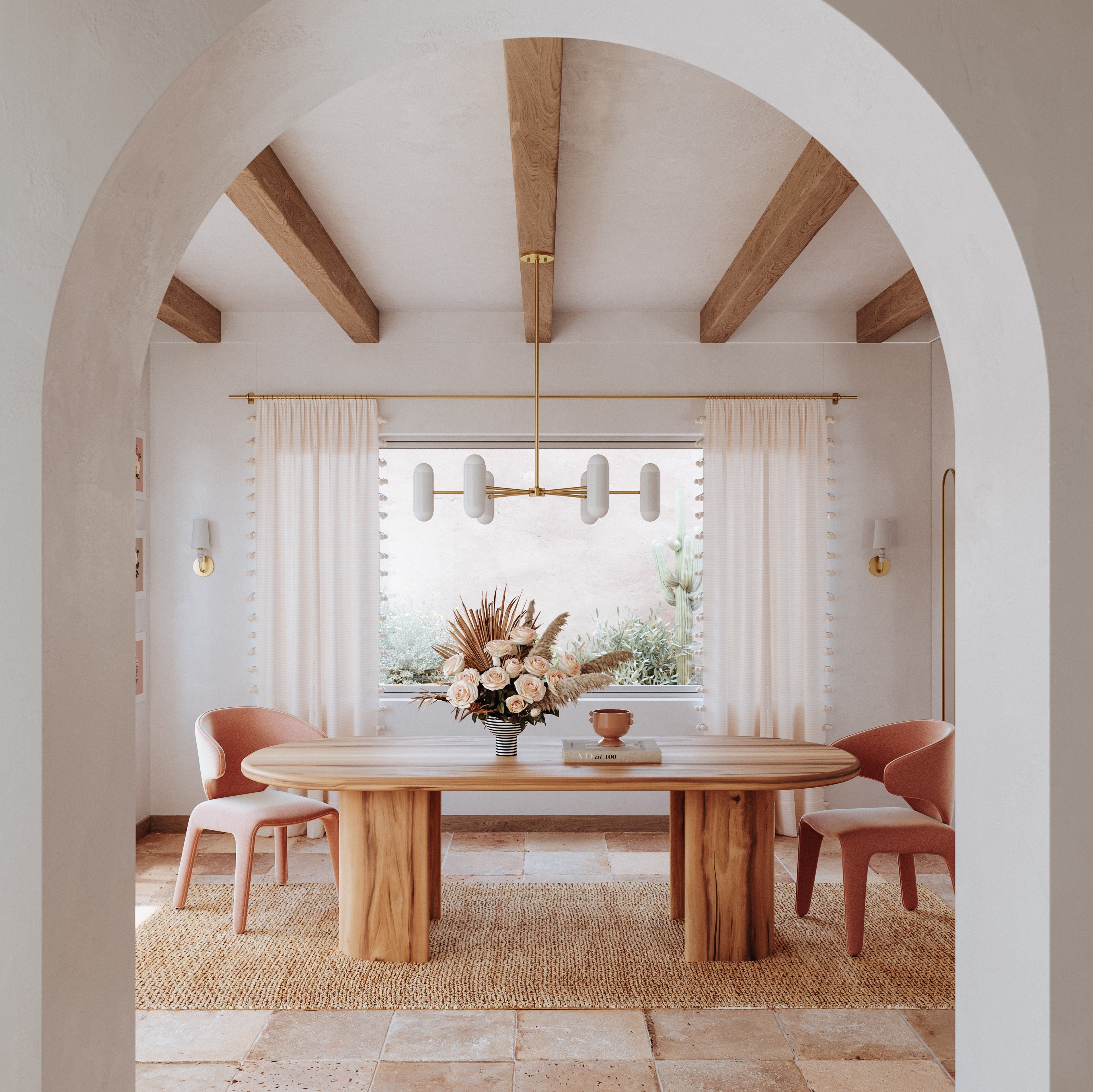 Dining room through a stucco arch with exposed wood beams, brass chandelier, soft curtains and wooden table with dried flower arrangement