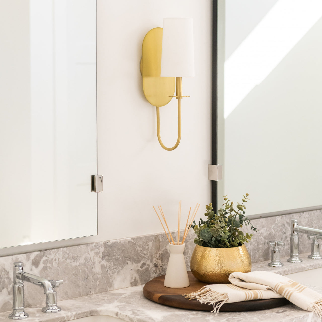 Gold wall sconce mounted beside a bathroom mirror above a marble vanity with a gold planter and reed diffuser