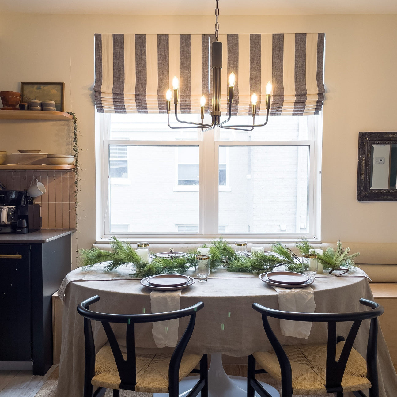 Dining table set with evergreen garland, plates, and candles beneath a lit black chandelier in a bright dining nook