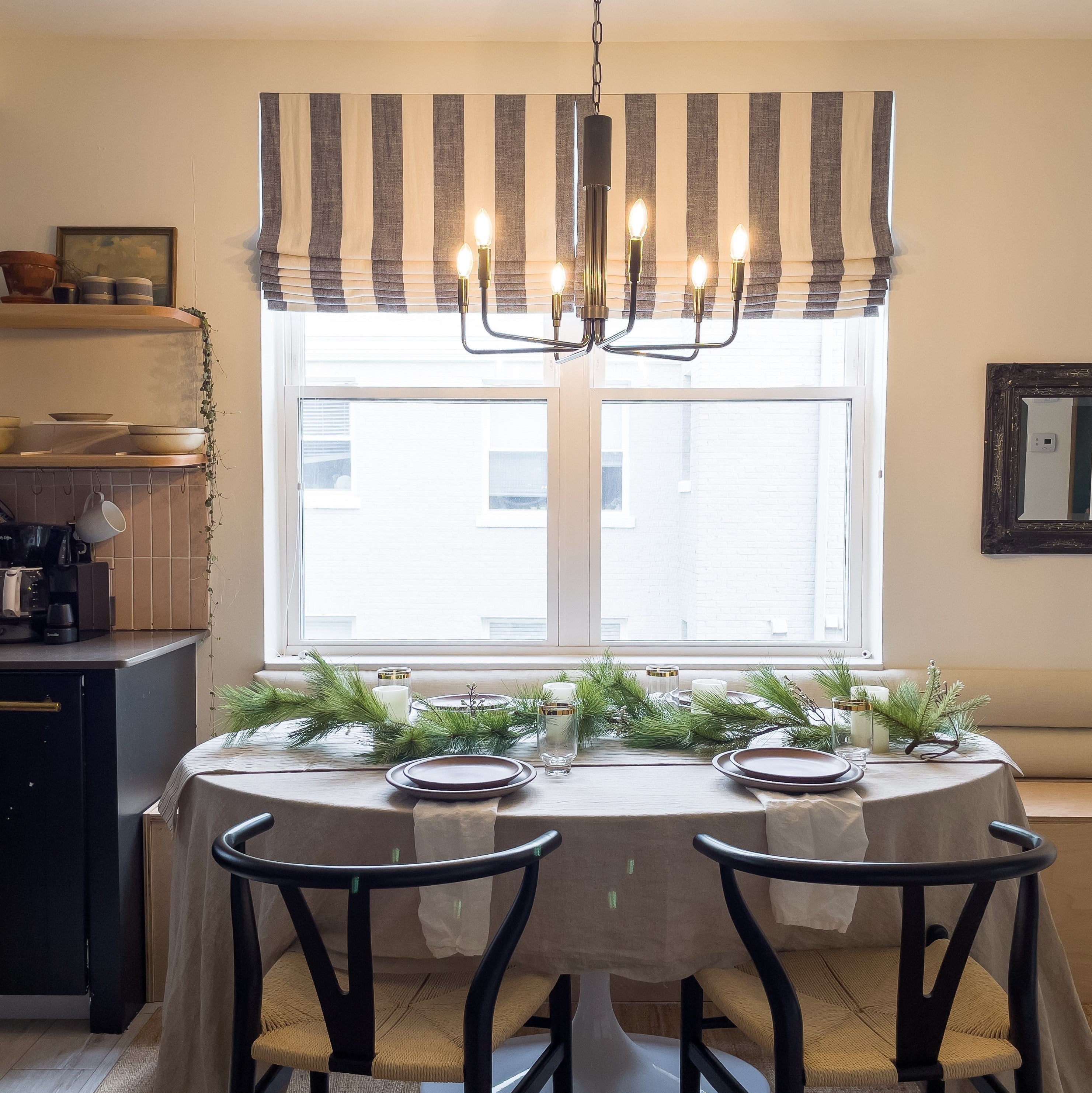 Dining table set with evergreen garland, plates, and candles beneath a lit black chandelier in a bright dining nook