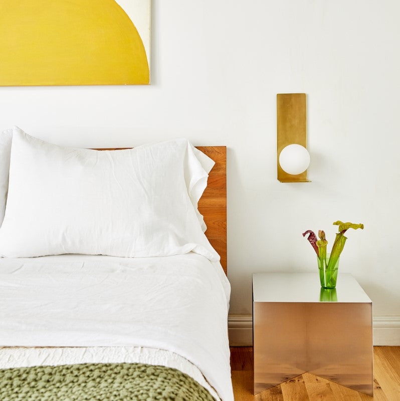 Bedside wall sconce over white bedding and wood headboard with mirrored nightstand and green vase in minimalist bedroom