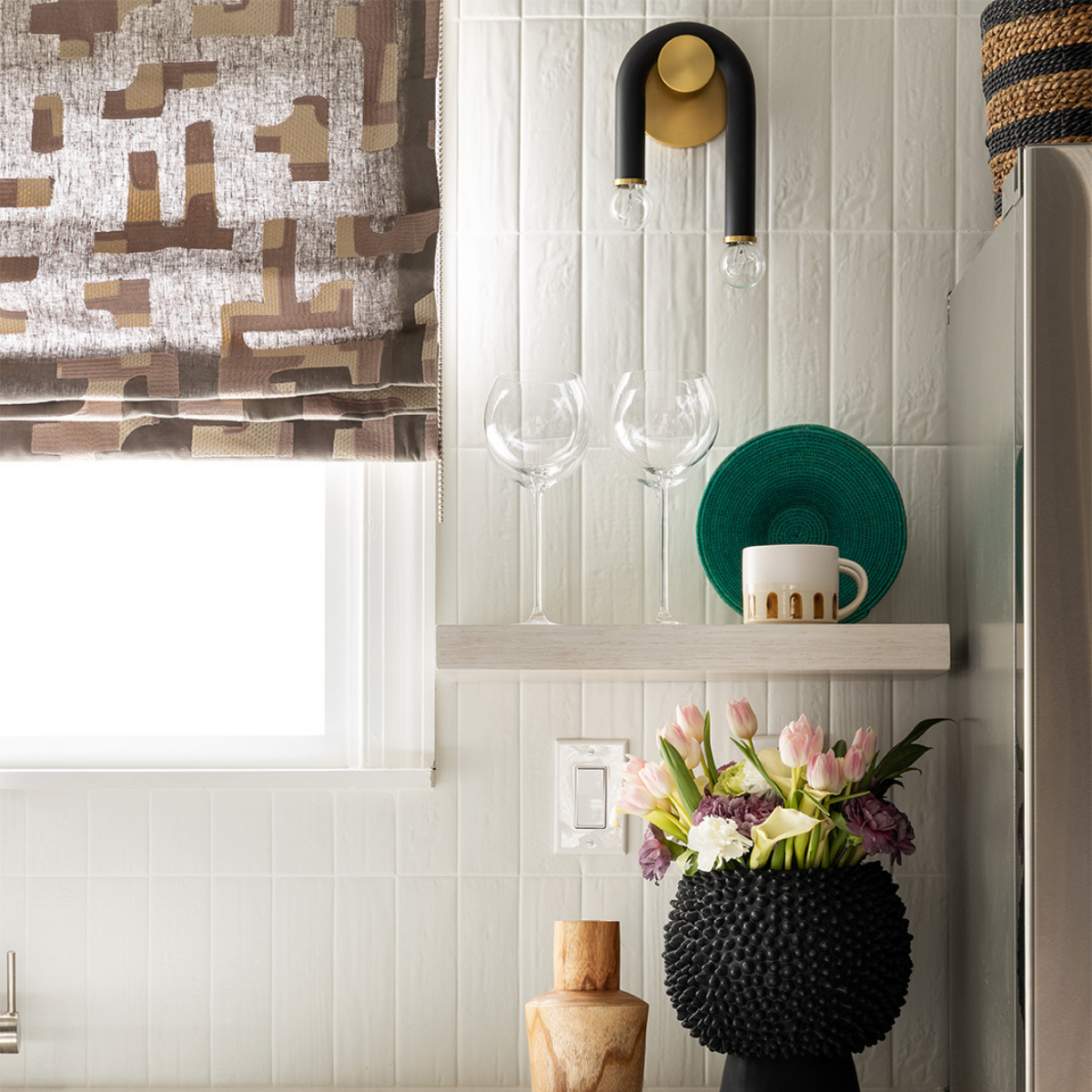 Black-and-gold U-shaped wall sconce over a shelf with two wine glasses, a green plate and mug, and a black textured vase of pink tulips