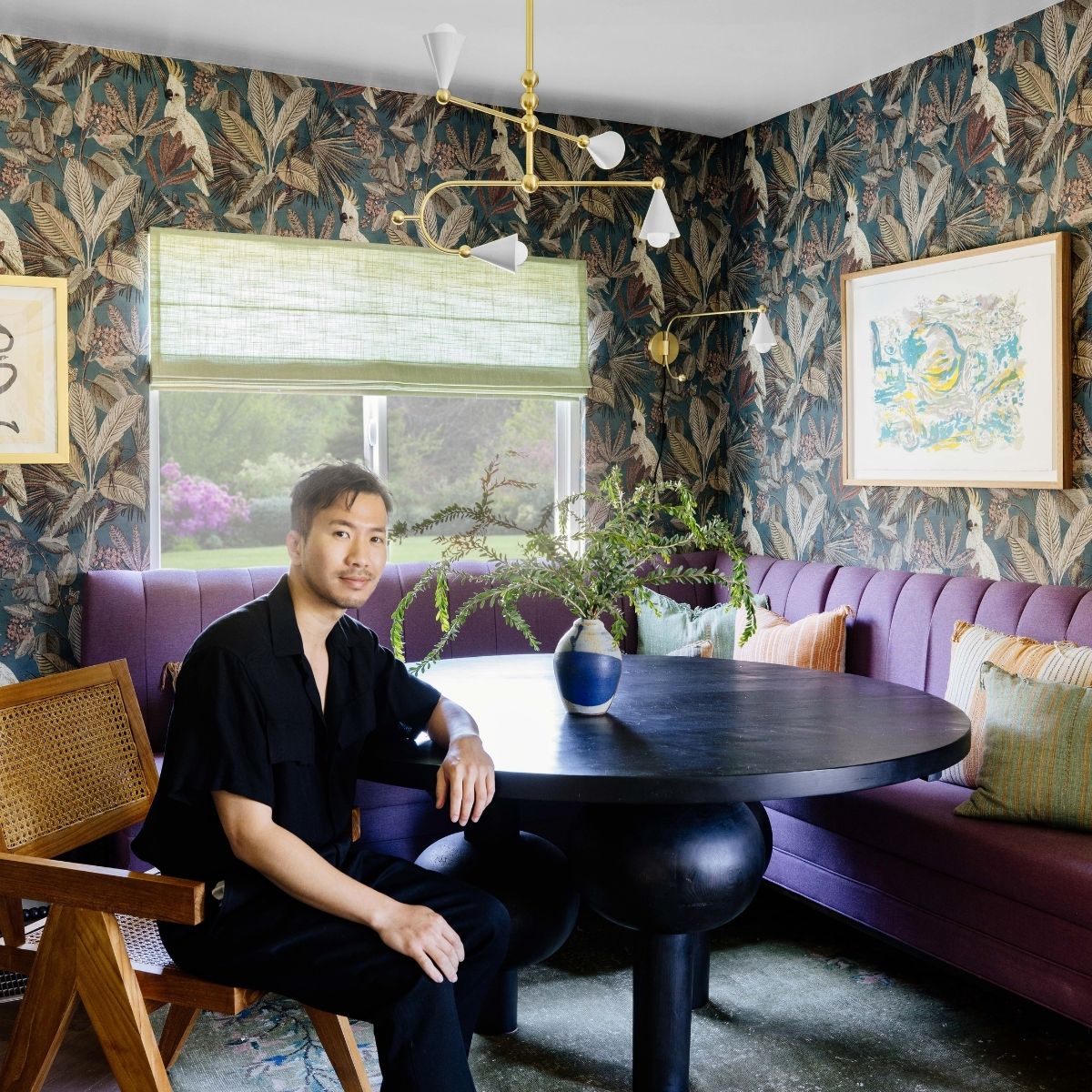Dabito seated at round black table in patterned-wallpaper nook with purple banquette, brass light fixture, and greenery