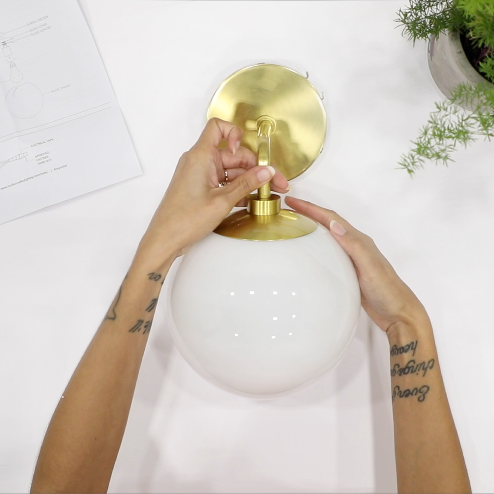 Hands attaching a white glass globe to a brass wall sconce from an overhead view on a white surface