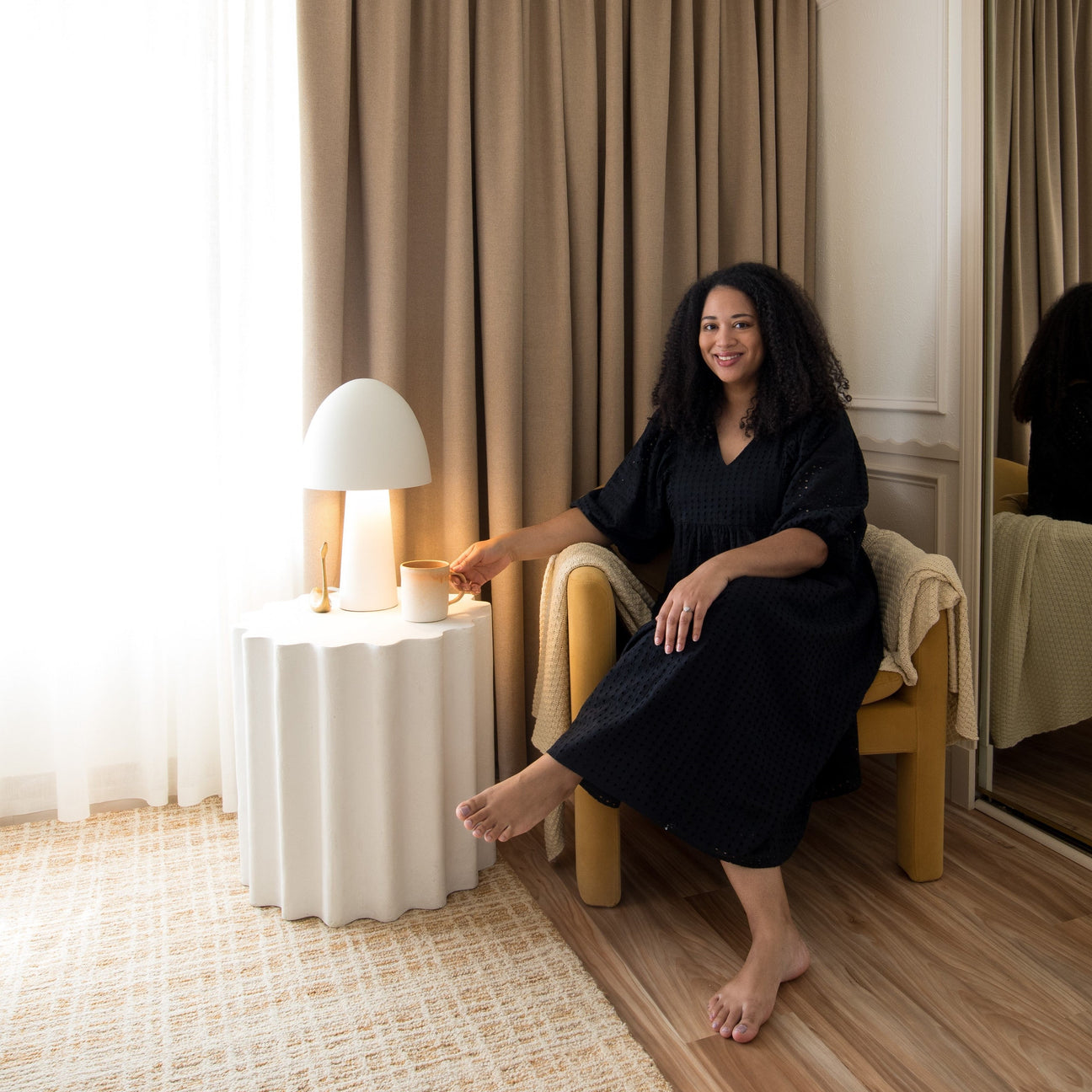 Amanda Walker sitting in a yellow armchair, smiling and holding a mug beside a white table lamp and tan curtains