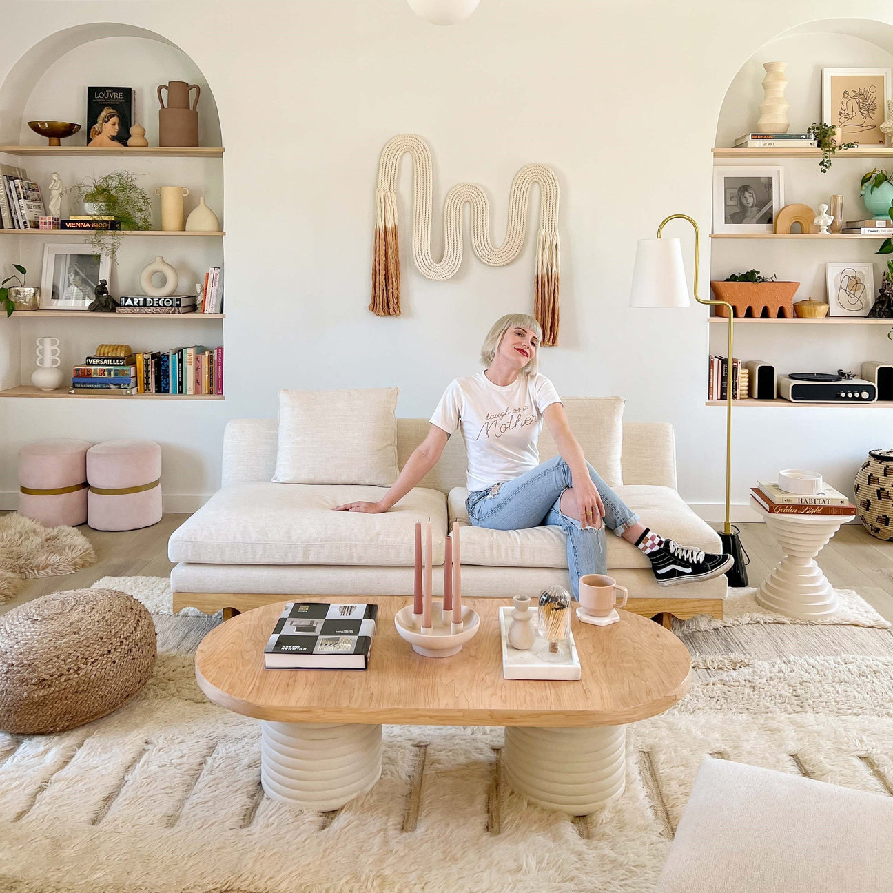 Woman sitting on a cream sofa smiling in a styled neutral living room with woven wall hanging and built-in shelves