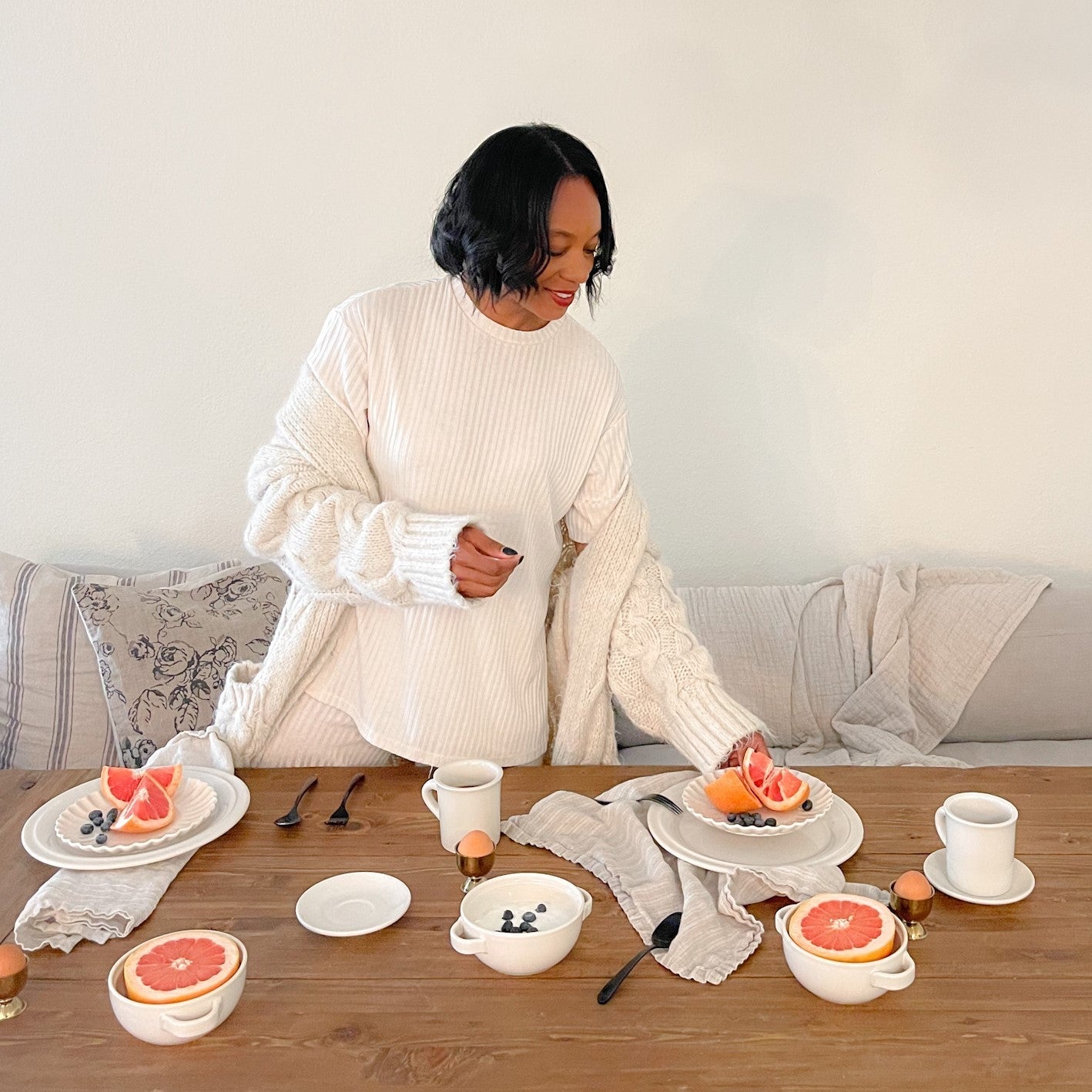 Ajai Guyot arranging grapefruit on a wooden breakfast table set with white dishes in a light neutral dining area