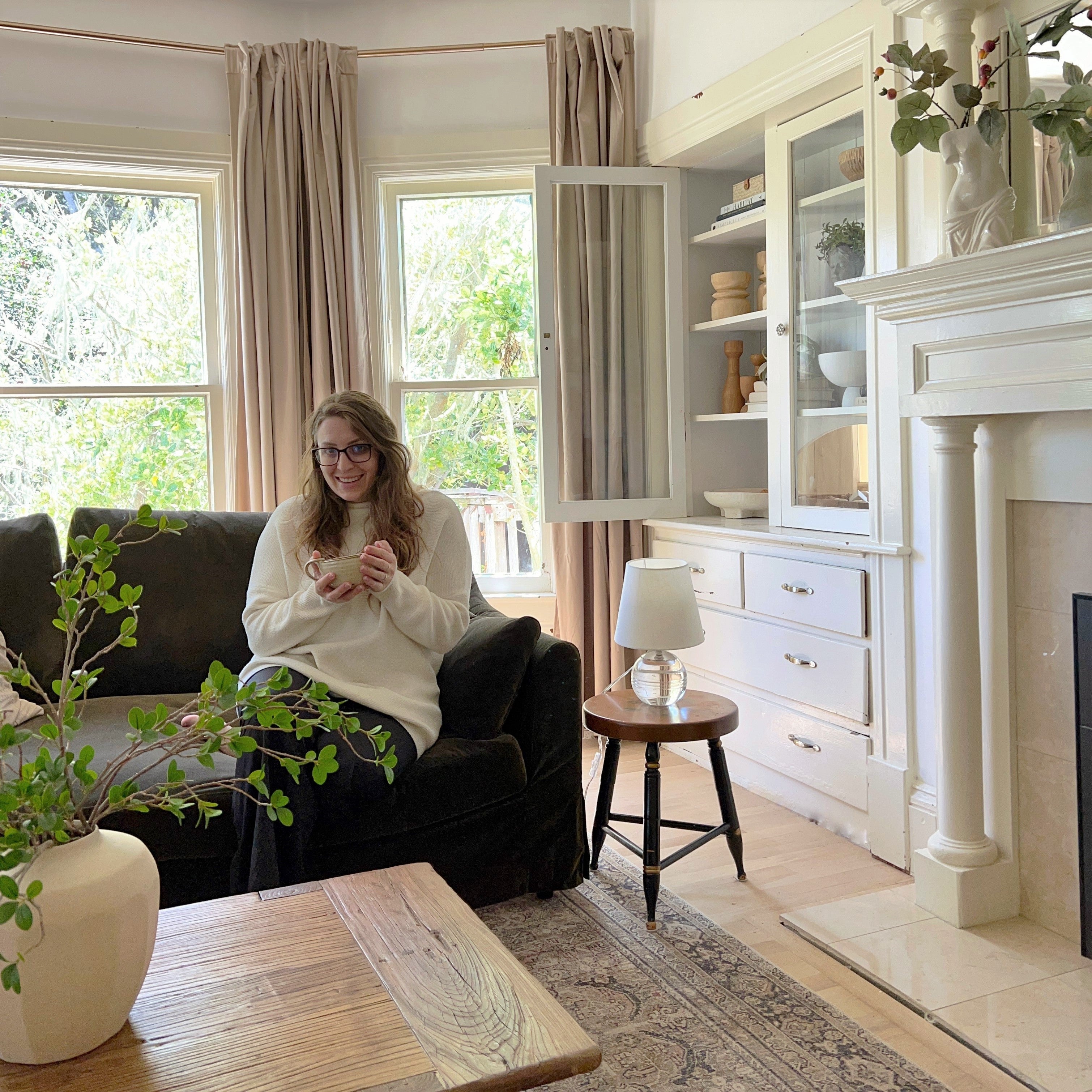 Erin Conway sitting on a dark sofa holding a mug in a bright living room with cream cabinets and plants