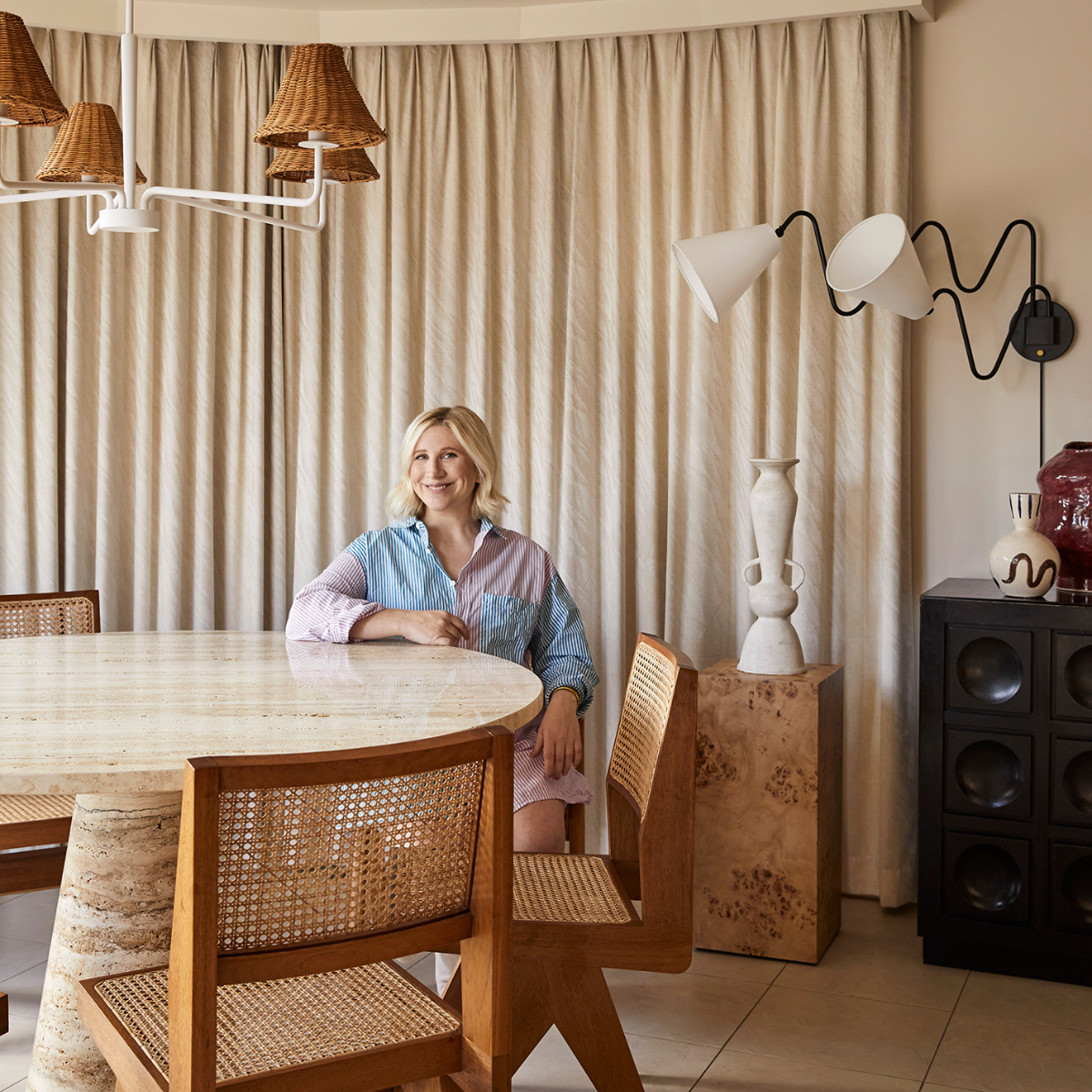 Woman seated at a round travertine dining table, smiling in a neutral dining room with cane chairs and sculptural wall lamp