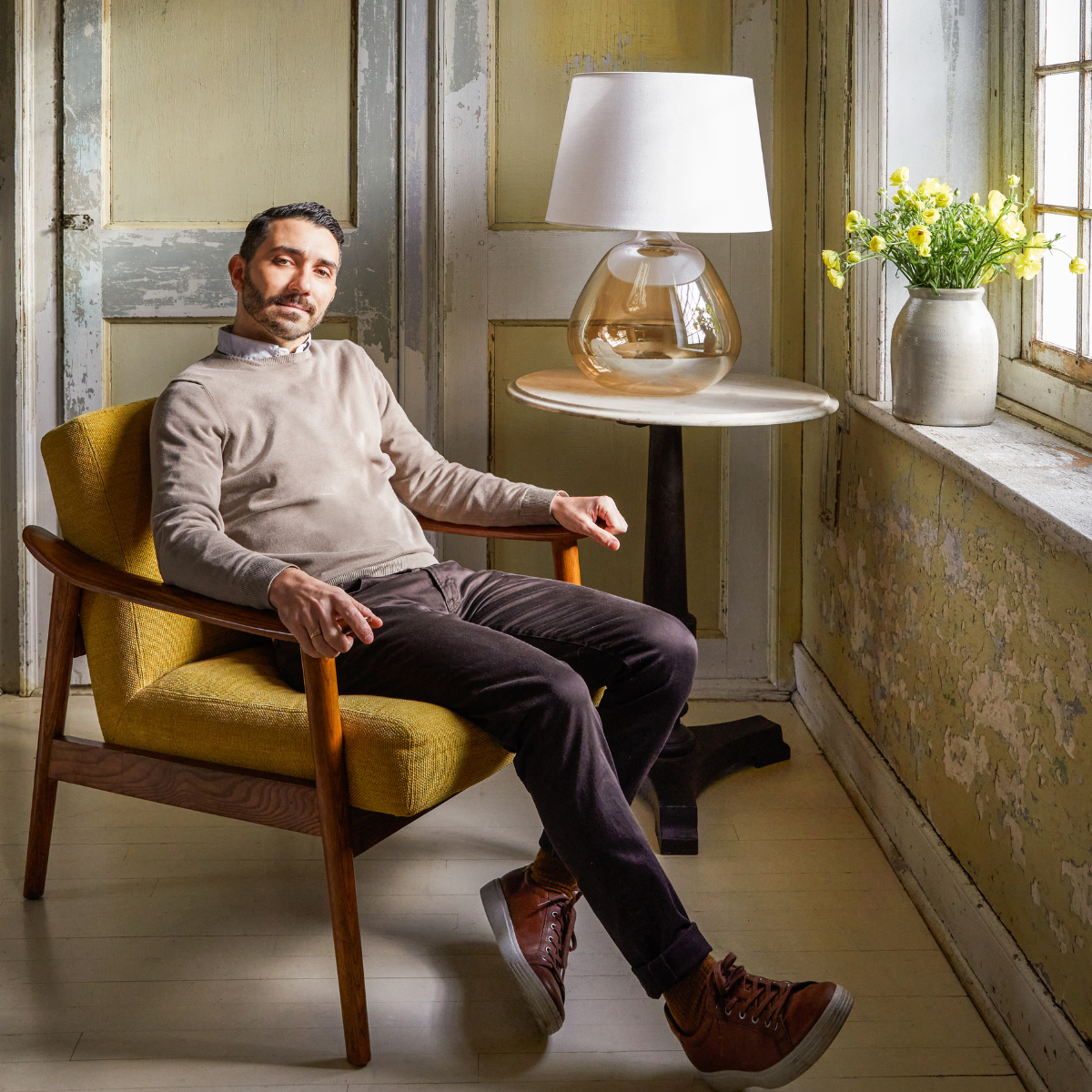 Man seated in mustard midcentury armchair next to round table with amber glass lamp and yellow flowers by sunlit window