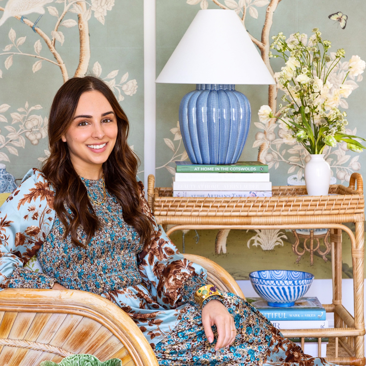 Smiling woman seated in a rattan chair beside a wicker side table with a blue ceramic lamp and vase of white flowers