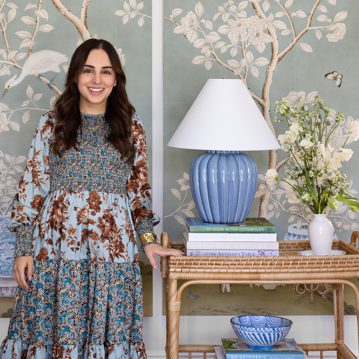 Woman smiling beside a blue ceramic table lamp on a wicker console with stacked books and a vase of white flowers against green floral wallpaper