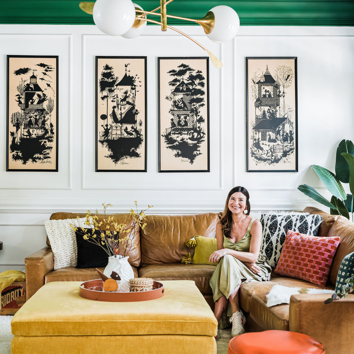 Woman seated on a tan leather sofa in a living room with black-and-beige prints, yellow ottoman, and brass globe chandelier