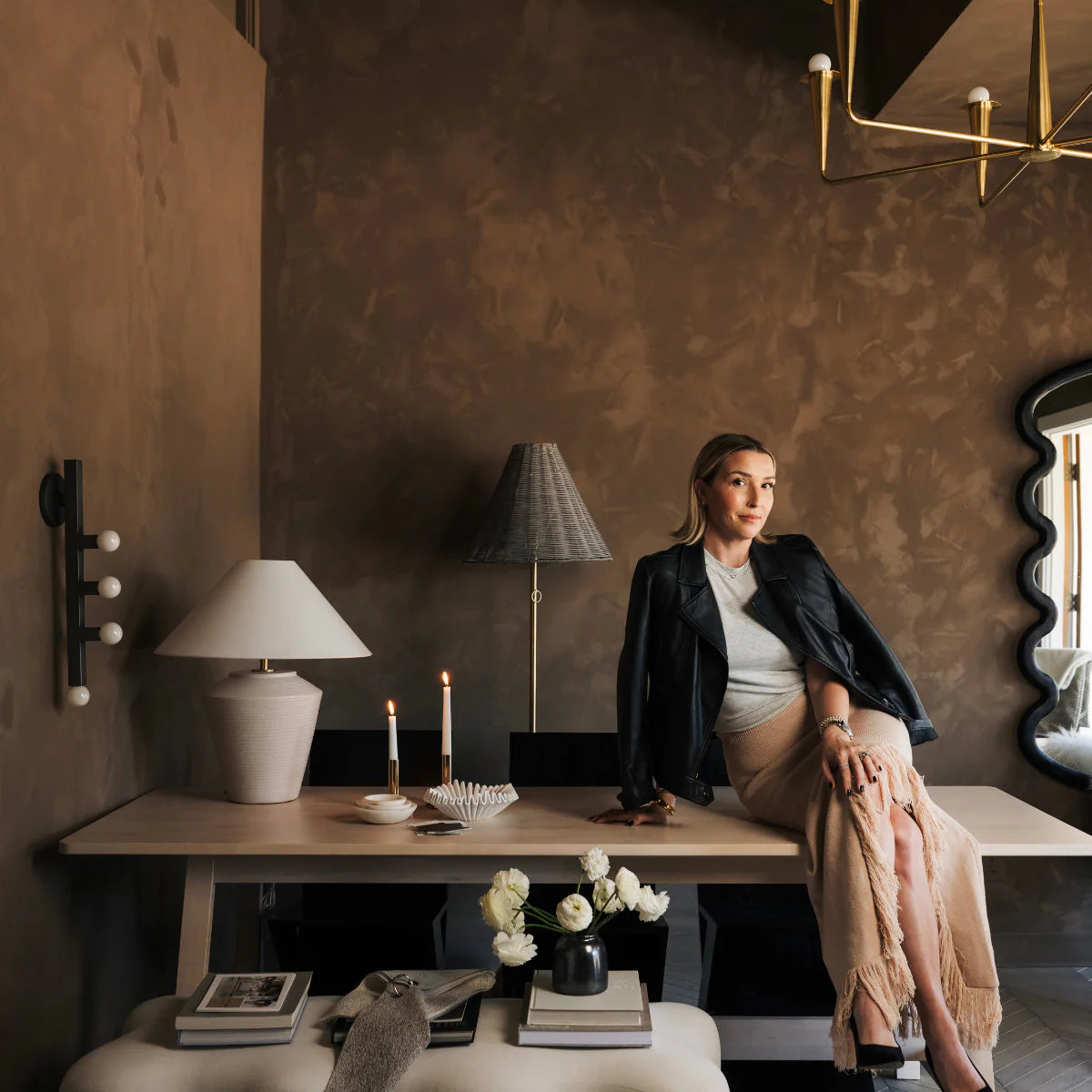 Woman seated on a desk wearing a leather jacket, surrounded by Mitzi lamps, candles, and decor in a warm brown interior