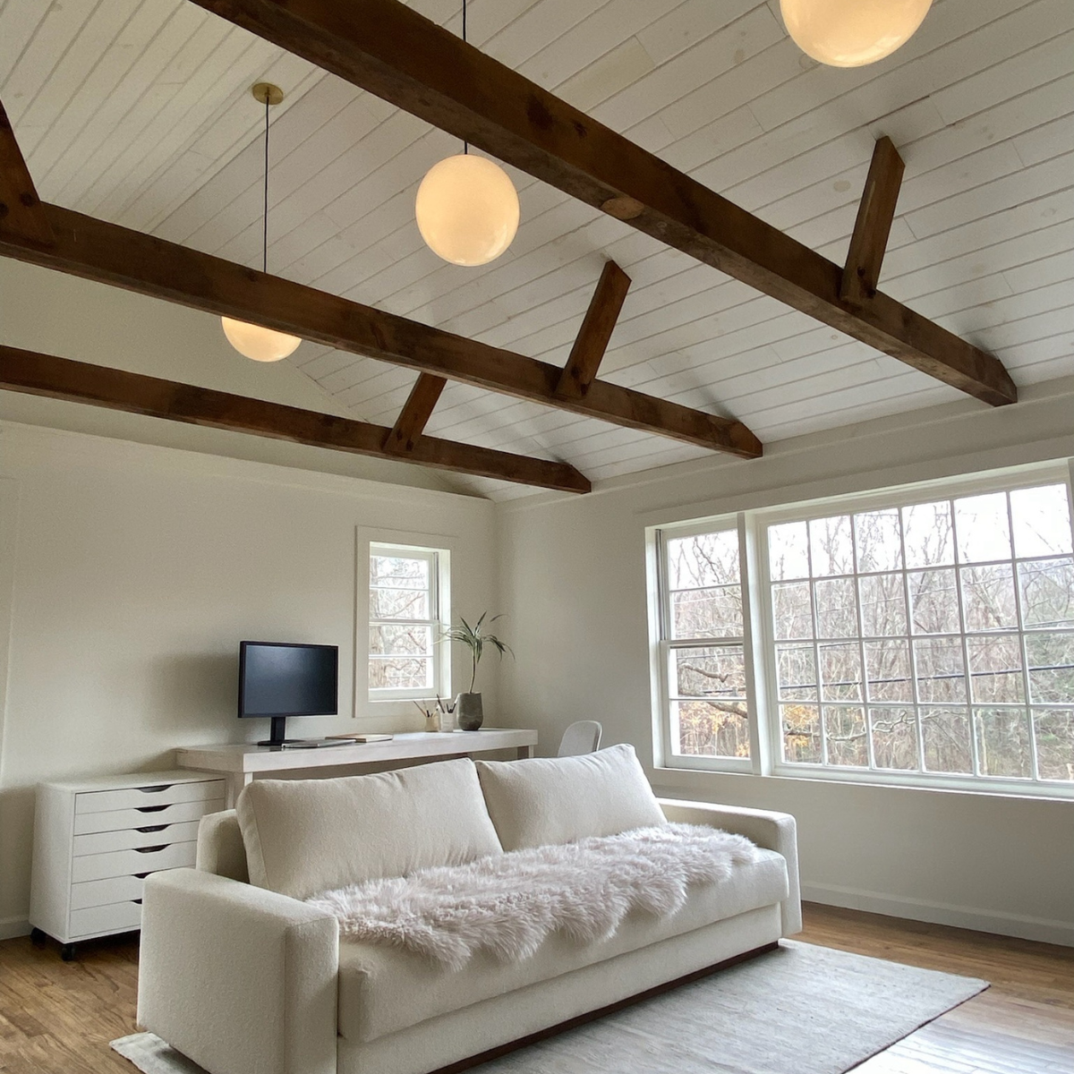 White sofa with faux-fur throw in bright living room under exposed wood beams and globe lights, windows with winter trees