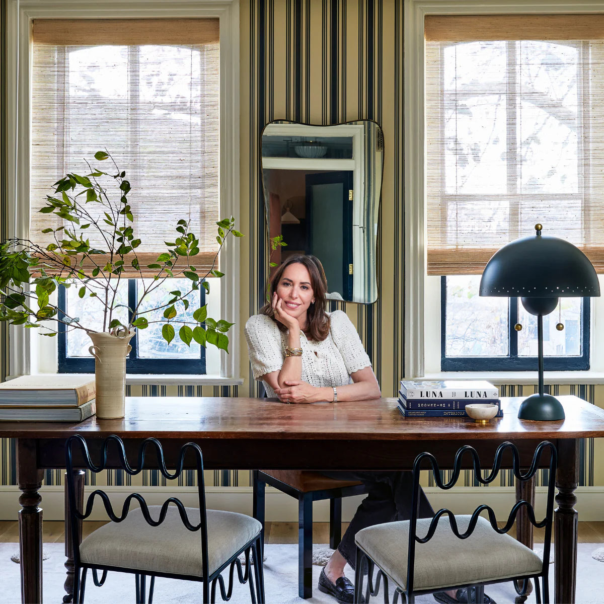 Woman seated at a wooden desk, leaning on her hand in a striped home office with a mirror, leafy vase, stacked books, and a black dome table lamp