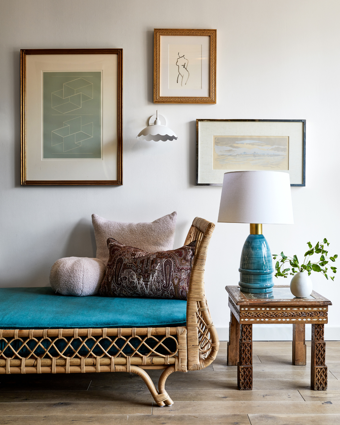 Rattan daybed with teal cushion and textured pillows beside carved wooden side table holding a blue ceramic lamp and vase