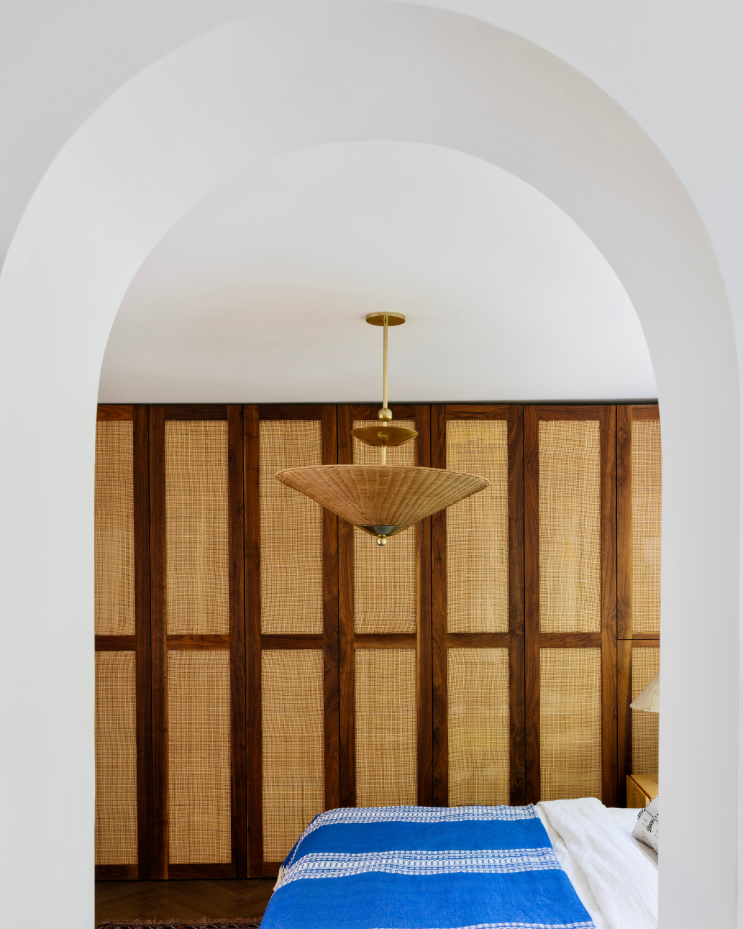 Woven rattan pendant light hanging over bed with blue blanket, seen through a white arched doorway and wood-and-cane wall