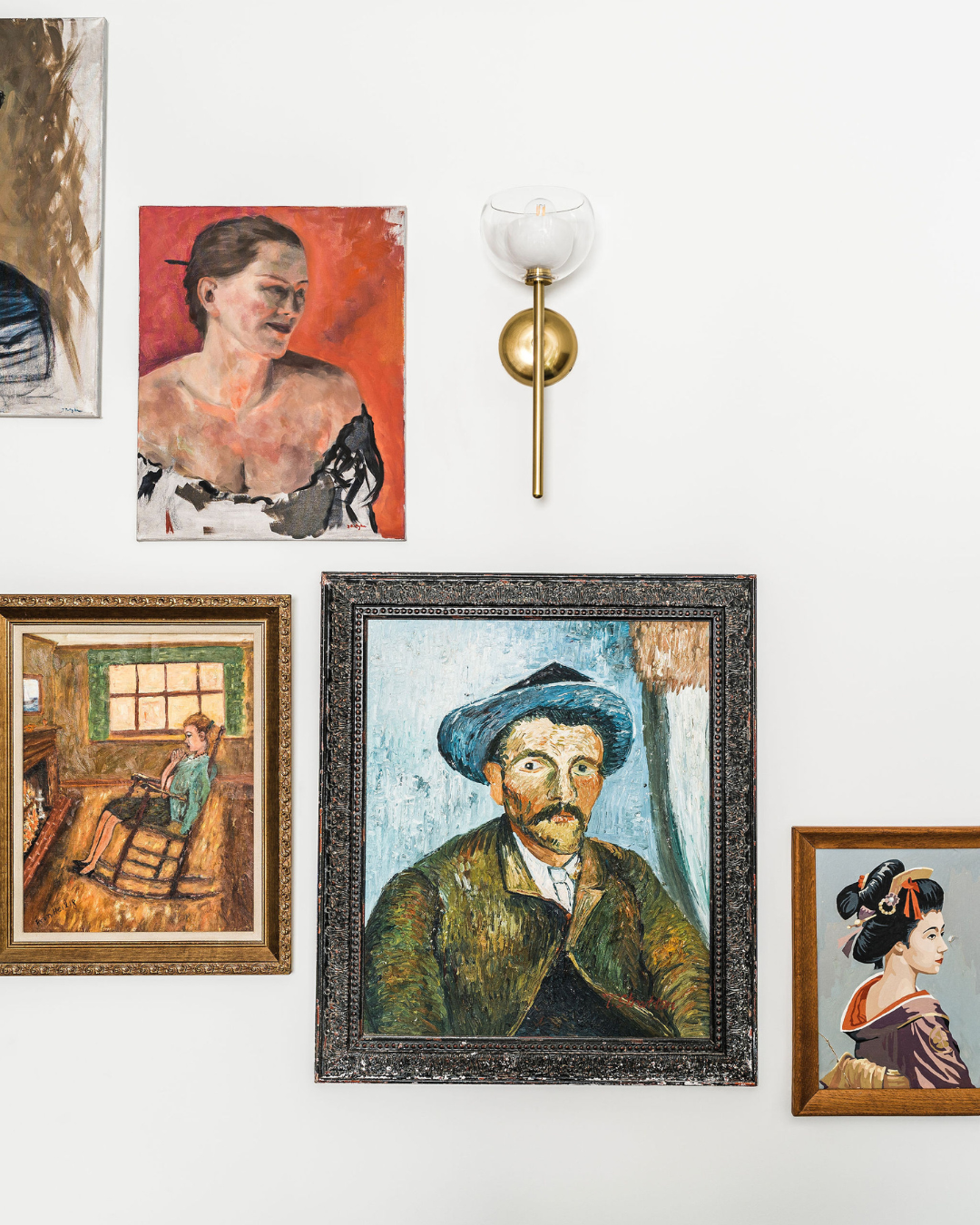 Gallery wall with multiple framed paintings and a brass and glass wall sconce on a white wall