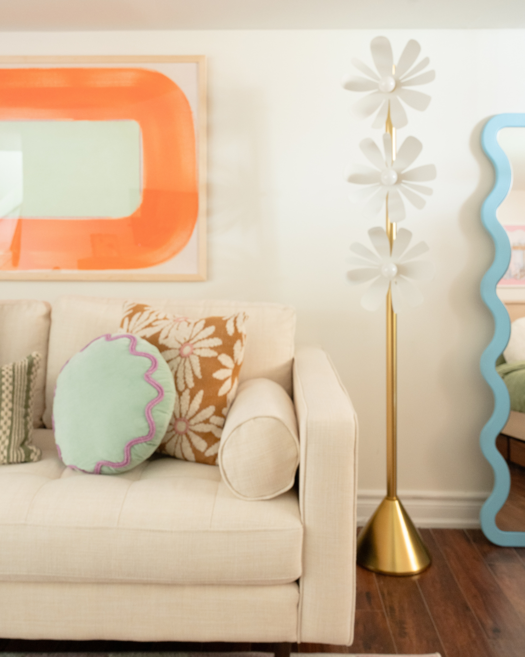 Beige sofa with mint round pillow and daisy patterned cushion beside a gold daisy floor lamp and orange abstract wall art