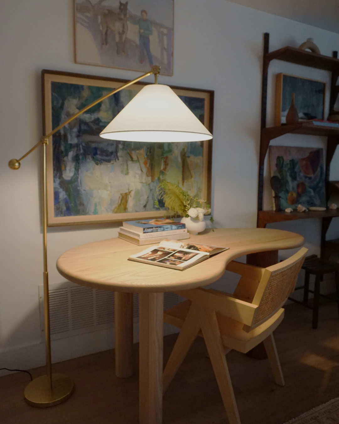 Brass floor lamp casting warm light over an oval wooden desk with an open book and cane-back chair in an art-filled corner