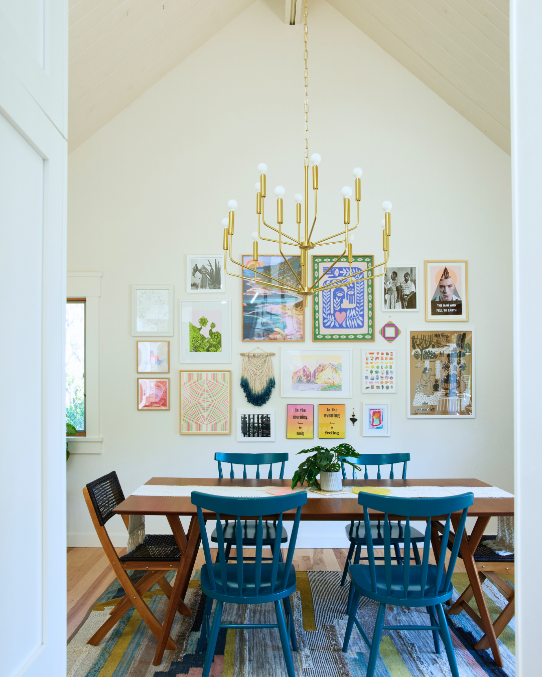 Dining table with teal chairs beneath a brass multi-arm chandelier in a bright vaulted room with a colorful gallery wall