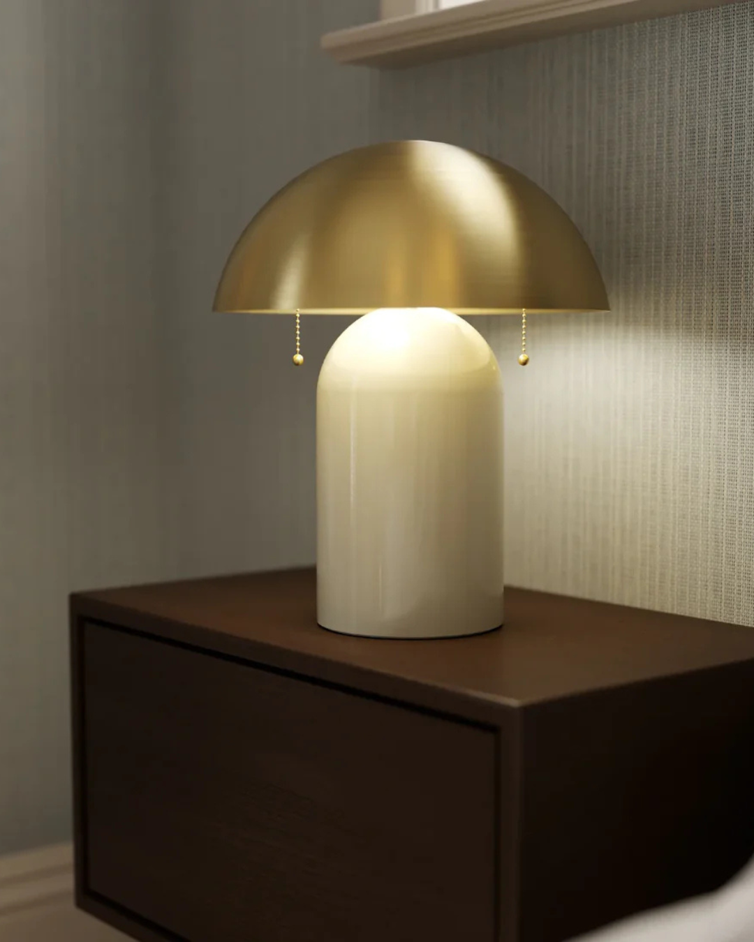Front view of lamp with brass dome shade and glossy ivory ceramic base on dark wood nightstand against gray textured wall