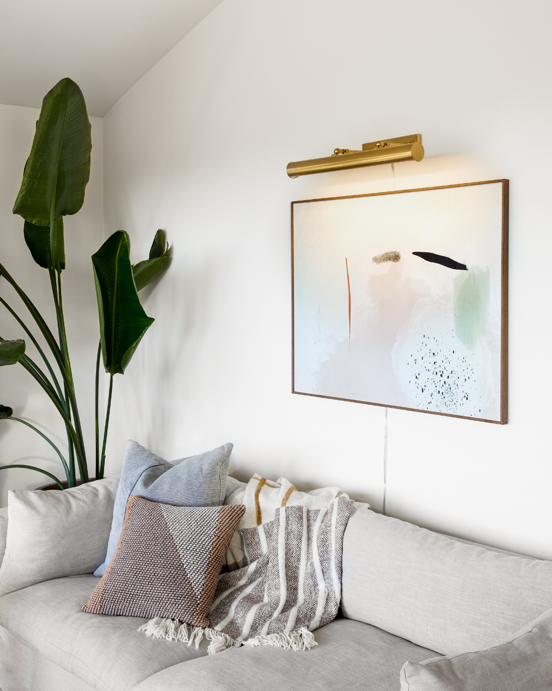 Cozy living room corner with light beige sofa, textured throw and patterned pillows beneath framed abstract art lit by a brass picture light and a tall potted plant