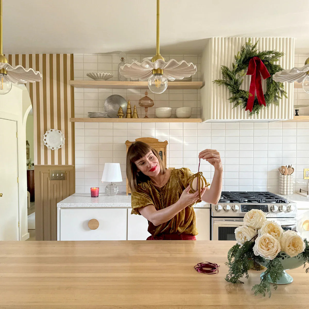Woman crafting a DIY ribbon ornament in a bright kitchen with tiled backsplash, wreath on the range hood, and white roses on the island