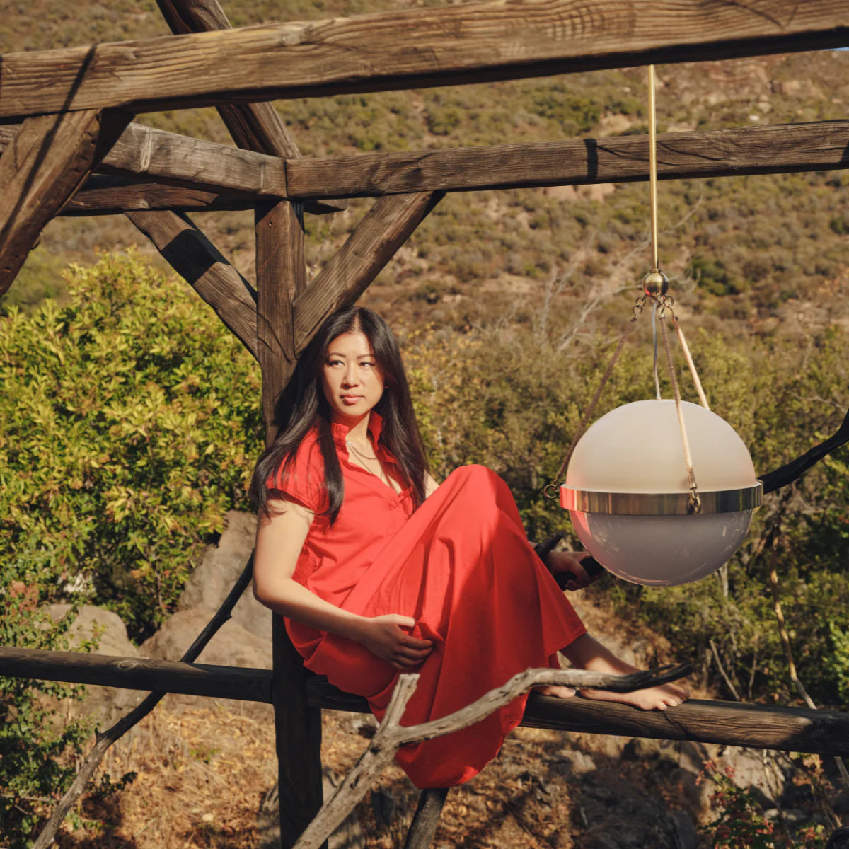 Woman in a red dress seated on a wooden pergola next to a hanging spherical pendant light with sunlit hillside in the background