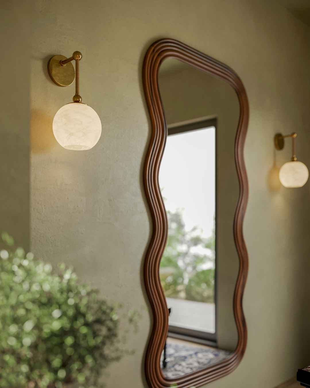 Curved wooden mirror and brass wall sconces beside a doorway in a softly lit entryway with a blurred potted plant in the foreground