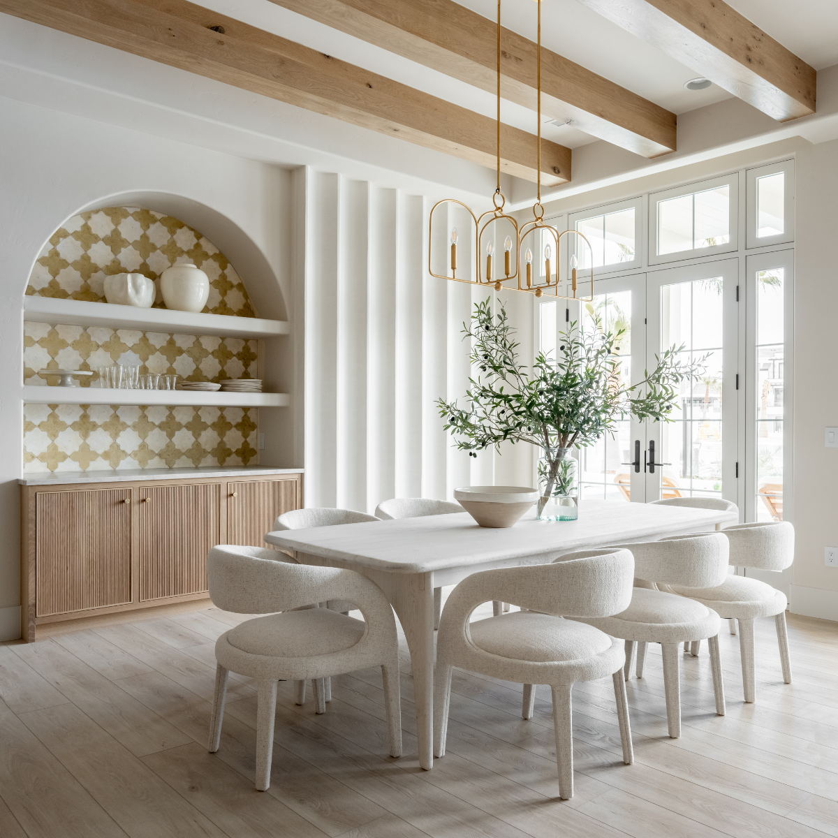 Dining table with six curved upholstered chairs under brass pendant lights in a bright organic‑modern dining room with arched shelving and exposed wood beams