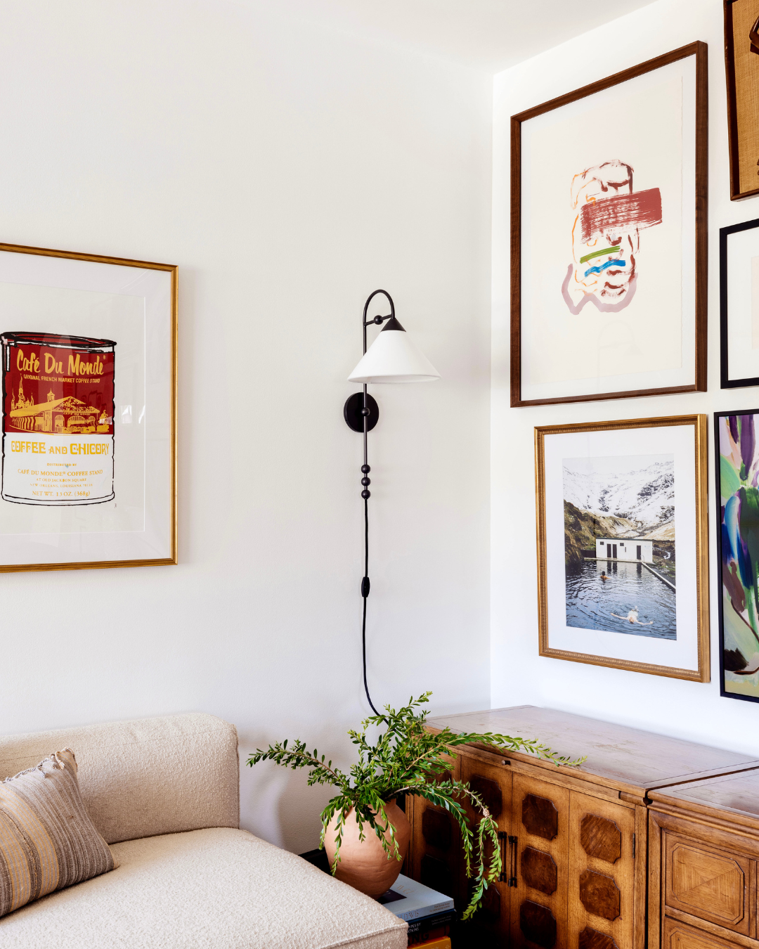 Corner of living room with cream sofa, black wall sconce, gallery wall including Cafe Du Monde poster, wooden sideboard and potted fern