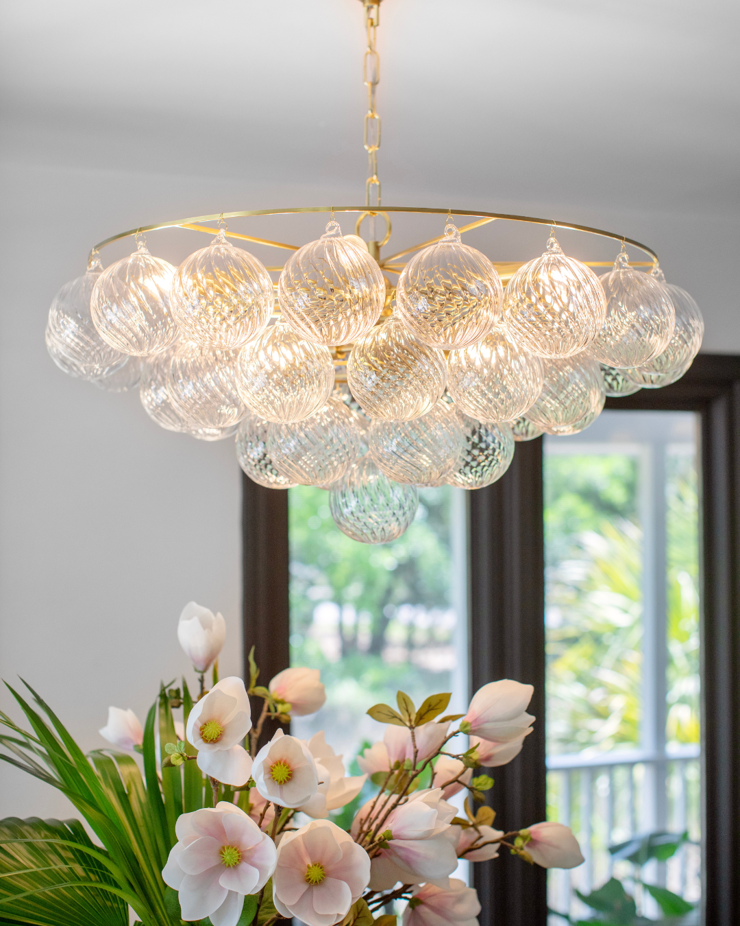 Front view of glass orb chandelier showcasing clustered ribbed glass spheres on brass ring with blurred window and flowers