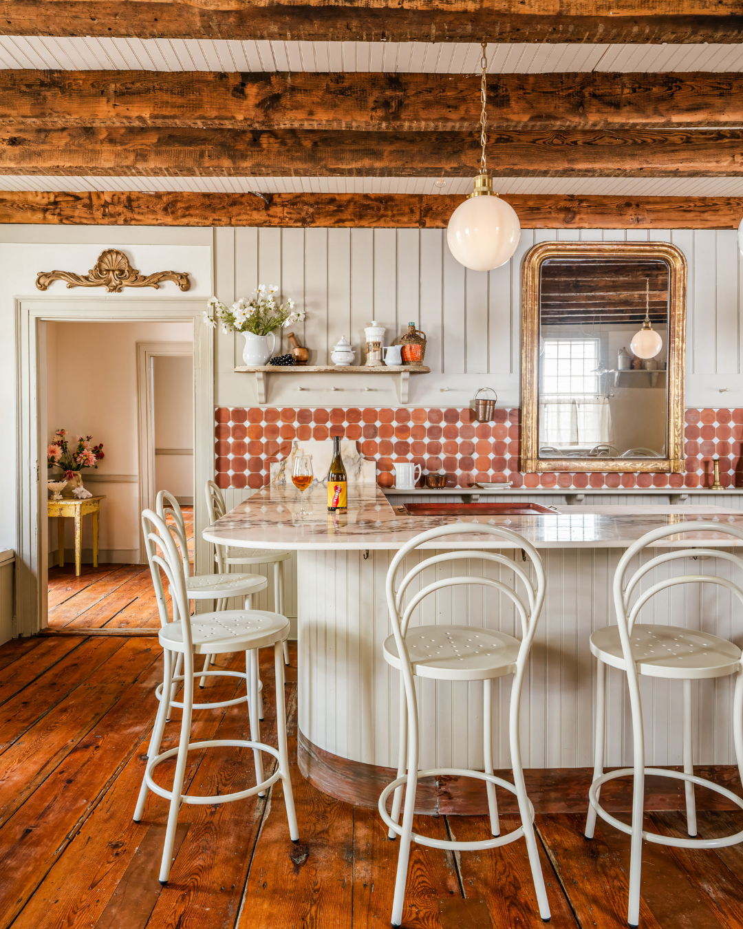 Sunlit country-style kitchen with marble-topped island, four white metal stools, terracotta hex tile backsplash, exposed wooden beams