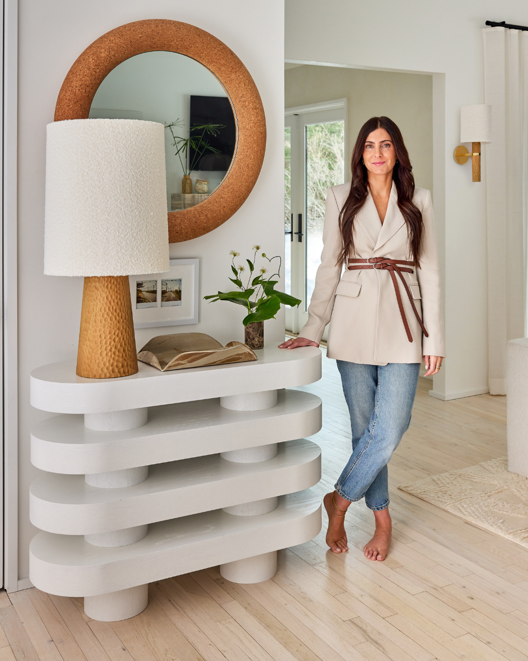 Woman standing barefoot beside white sculptural console table with gold lamp and round cork mirror in bright room